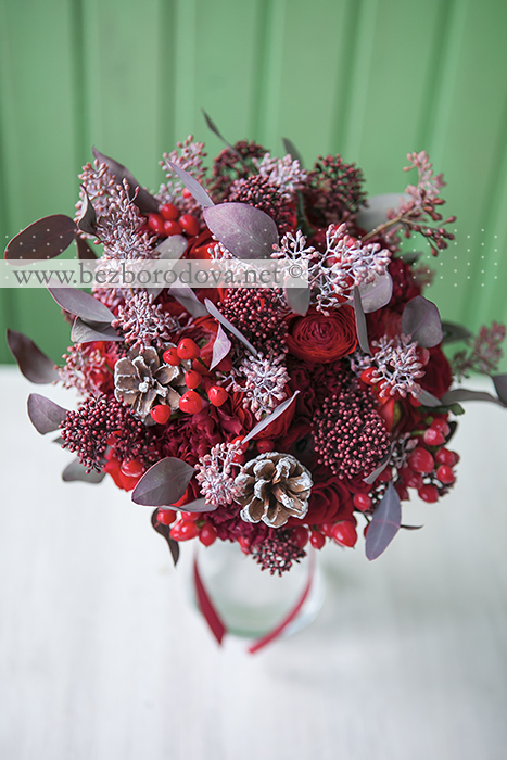 Зимний свадебный букет из красных роз и ранункулюсов с ягодами гиперикума и шишками 